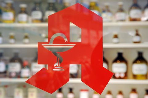 Auf einer Glasscheibe klebt das rote Apotheken-"A", unscharf im Hintergrund braune Apothekerflaschen in einem Regal