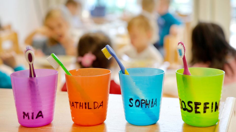 Vier bunte, mit Namen beschriftete Zahnputzbecher stehen auf einem Regal. Unscharf im Hintergrund sind Kinder zu erahnen