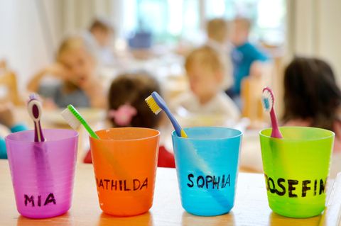 Vier bunte, mit Namen beschriftete Zahnputzbecher stehen auf einem Regal. Unscharf im Hintergrund sind Kinder zu erahnen