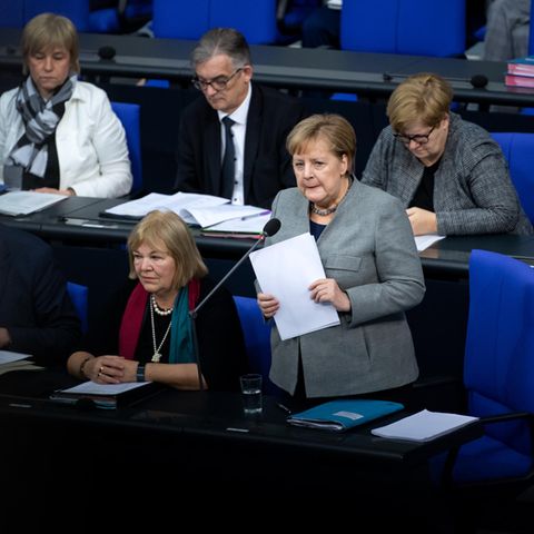 An ihrem Sitzplatz im Bundestag steht Angela Merkel, hält Papiere in den Händen und spricht in ein Mikrofon