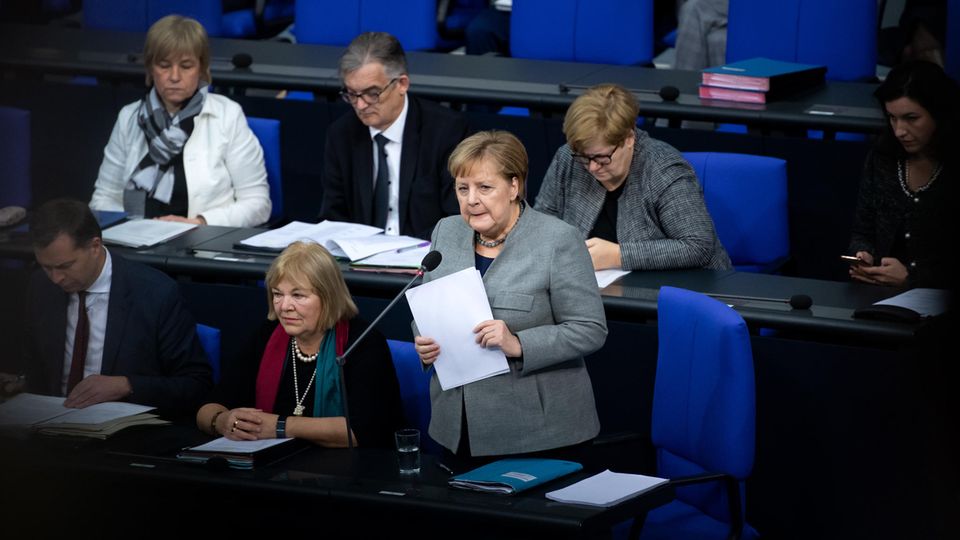 An ihrem Sitzplatz im Bundestag steht Angela Merkel, hält Papiere in den Händen und spricht in ein Mikrofon