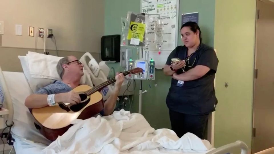 Krankenschwester steht am Bett und singt mit einem Patienten, der Gitarre spielt