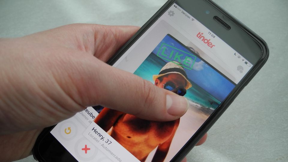 Ein Smartphone-Display zeigt das Tinder-Profilfoto eines Mannes mit nacktem Oberkörper am Strand