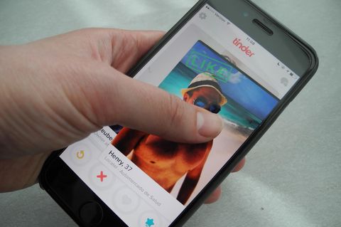 Ein Smartphone-Display zeigt das Tinder-Profilfoto eines Mannes mit nacktem Oberkörper am Strand