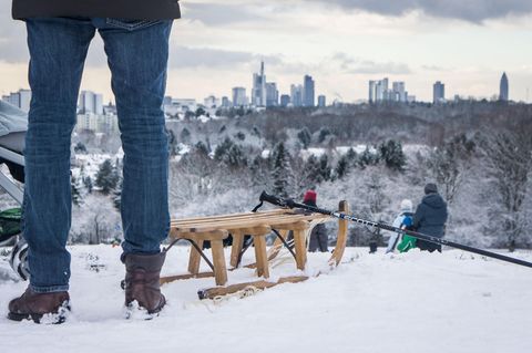 Neben einem Holzschlitten sind die Beine eines Mannes zu sehen. Im Hintergrund die Skyline von Frankfurt am Main