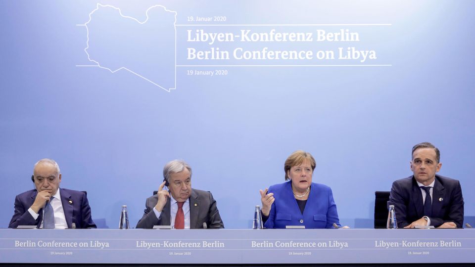 Auf einem Podium mit Bundesadler sitzen drei Männer in Anzügen und Bundeskanzlerin Angela Merkel. Sie trägt einen blauen Blazer
