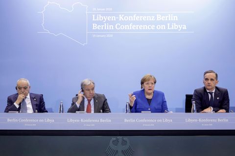 Auf einem Podium mit Bundesadler sitzen drei Männer in Anzügen und Bundeskanzlerin Angela Merkel. Sie trägt einen blauen Blazer