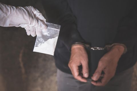 Ein Drogendealer wird verhaftet und trägt Handschellen. Eine Hand im Handschuh hält eine Tüte mit weißem Pulver hoch.