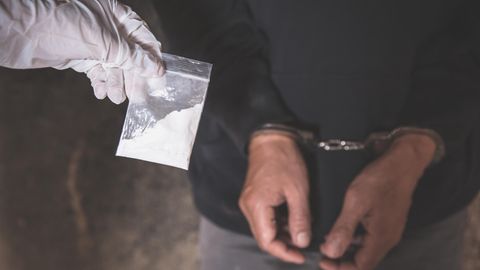 Ein Drogendealer wird verhaftet und trägt Handschellen. Eine Hand im Handschuh hält eine Tüte mit weißem Pulver hoch.
