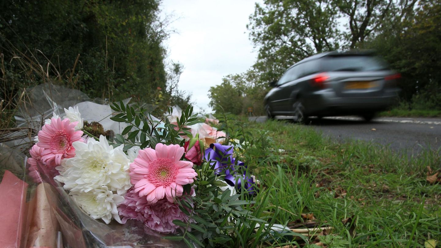 Ein Blumenstrauß mit rosa Gerbera und weißen Nelken liegt am Straßenrand, während ein graues Auto schnell vorbeifährt