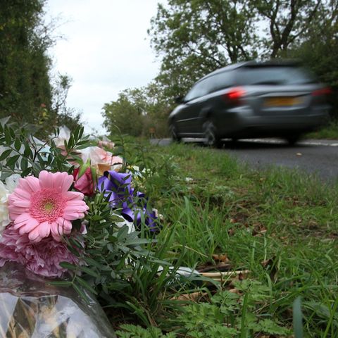 Ein Blumenstrauß mit rosa Gerbera und weißen Nelken liegt am Straßenrand, während ein graues Auto schnell vorbeifährt
