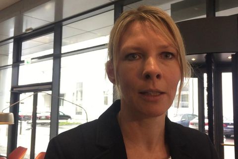 Eine blonde Frau mit schwarzem Blazer spricht in die Kamera. Im Hintergrund eine Glasfront mit Drehtür