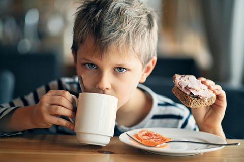 Ein Junge trinkt aus einer Tasse und hält ein Schinkenbrot in der Hand