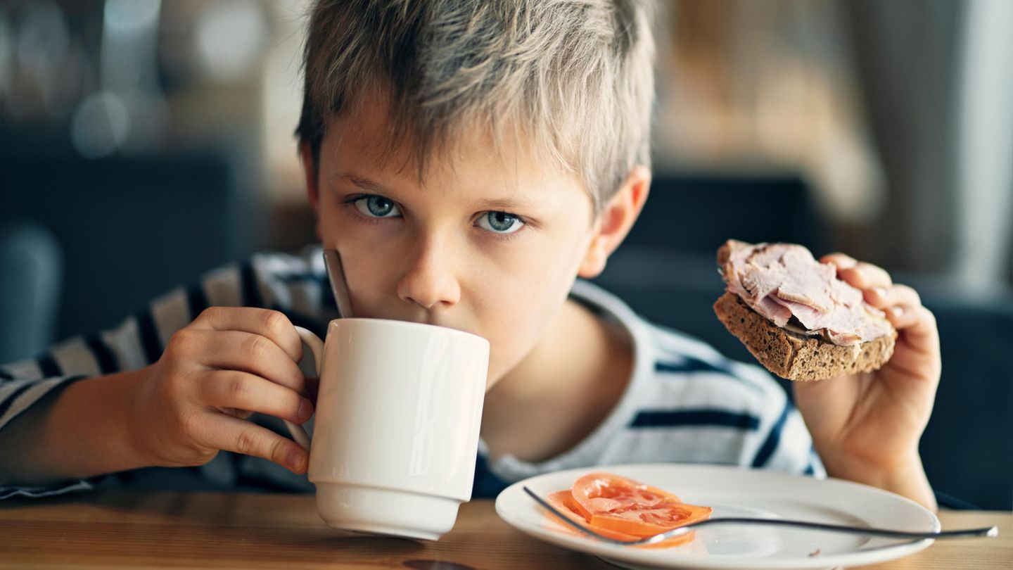 Ein Junge trinkt aus einer Tasse und hält ein Schinkenbrot in der Hand