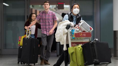 Am Flughafen: Eine Frau trägt eine Atemmaske neben ankommenden Reisenden ohne Mundschutz