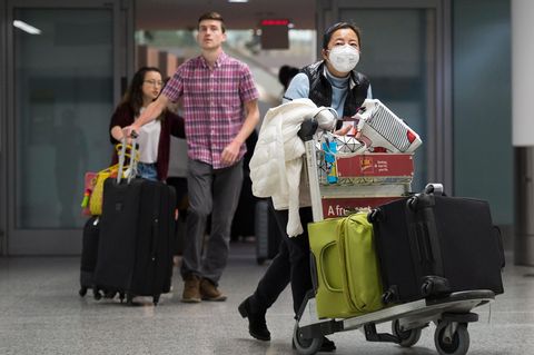 Am Flughafen: Eine Frau trägt eine Atemmaske neben ankommenden Reisenden ohne Mundschutz