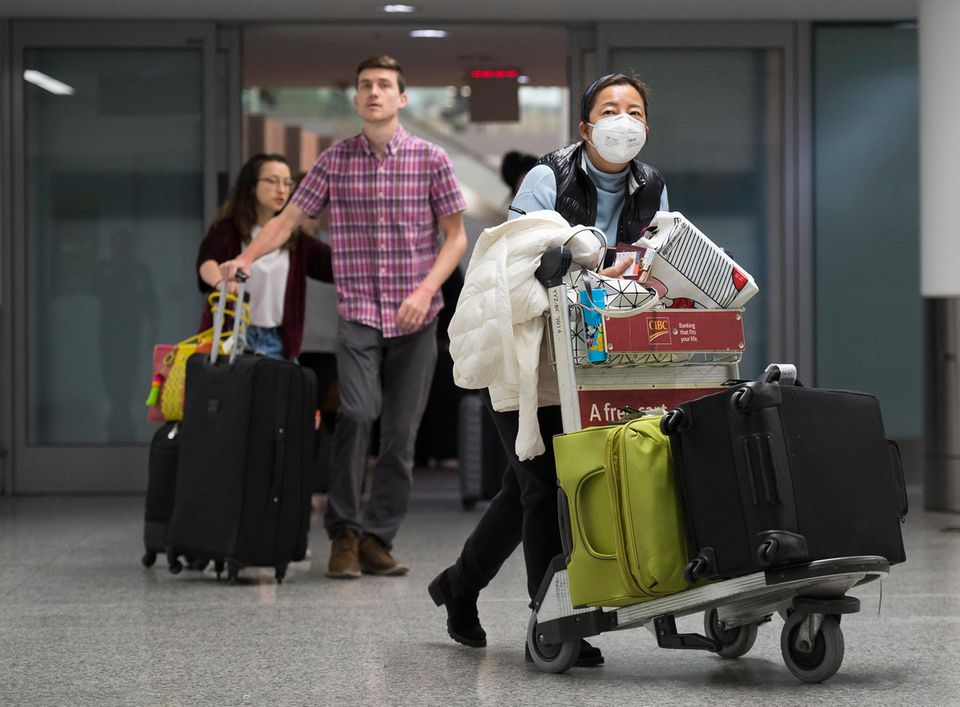 Fragen und Antworten: Am Flughafen: Eine Frau trägt eine Atemmaske neben ankommenden Reisenden ohne Mundschutz Am Flughafen: Eine Frau trägt eine Atemmaske neben ankommenden Reisenden ohne Mundschutz