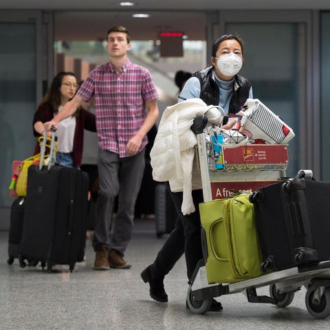 Am Flughafen: Eine Frau trägt eine Atemmaske neben ankommenden Reisenden ohne Mundschutz