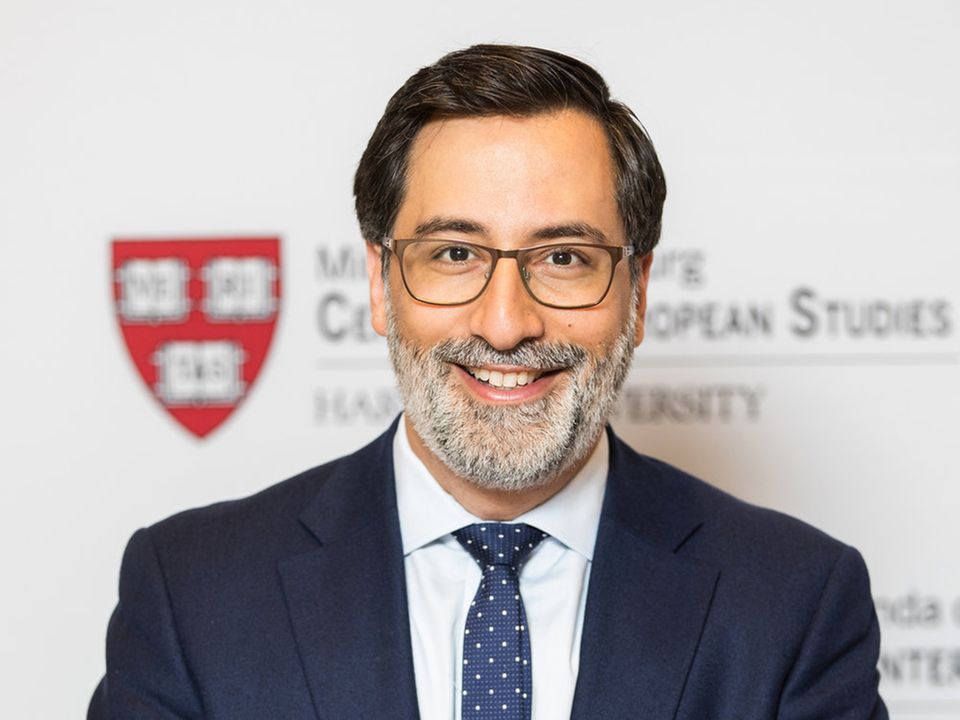 Prof. Dr. Dr. Alexander Görlach Ein Mann mit Brille, schwarzem Seitenscheitel und grauem Vollbart sitzt im Anzug vor einer Wand mit dem Logo der Uni Harvard