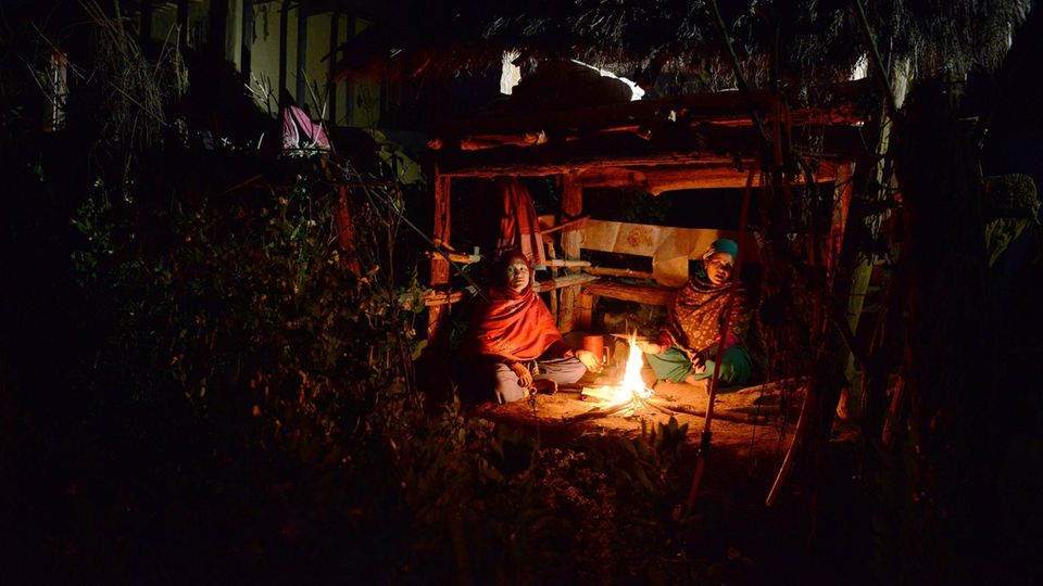 Menstruationshütte Zwei Frauen in bunten Gewändern sitzen nachts an einem Feuer vor einer einfachen Hütte