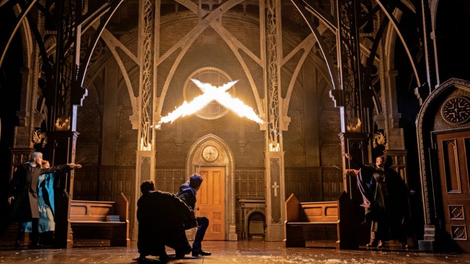 Harry Potter und das verwunsche Kind Theater Zwei Zauberer knien mit gezückten Zauberstäben vor zwei Säulen, aus denen Flammen schlagen. Im Hintergrund eine Holztür