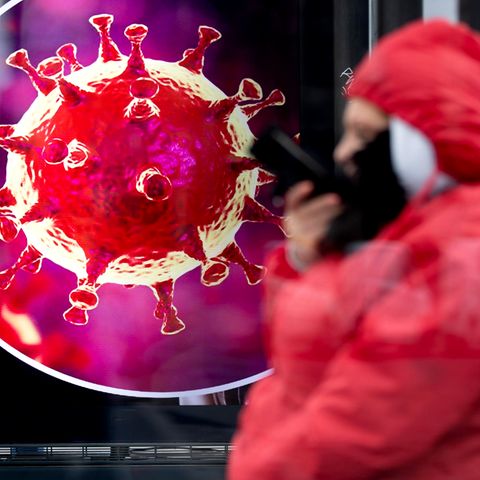 Eine Frau sitzt an einer Bushaltestelle vor einer elektronischen Werbetafel mit einer Illustration des Coronavirus.