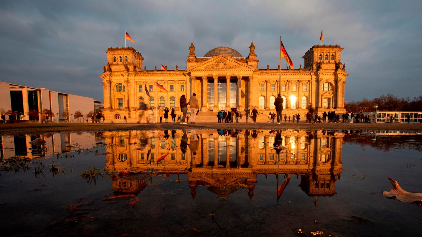 Das Reichstagsgebäude in Berlin liegt vor einem grauen Himmel in der Abendsonne und spiegelt sich im Wasserbecken davor