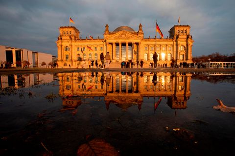 Das Reichstagsgebäude in Berlin liegt vor einem grauen Himmel in der Abendsonne und spiegelt sich im Wasserbecken davor