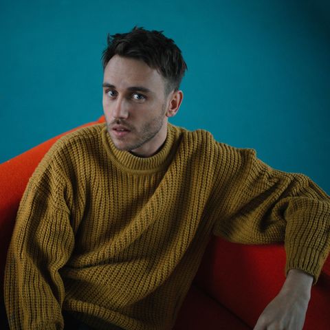 Clueso im gelben Strickpullover auf einem roten Sessel mit blauem Hintergrund
