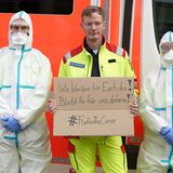 #FlattenTheCurve: Ein Notarzt hält, eingerahmt von zwei Rettungssanitätern in Infektionsschutzkleidung, ein Schild mit der Aufschrift "Wir bleiben für euch da! Bleibt für uns daheim!".