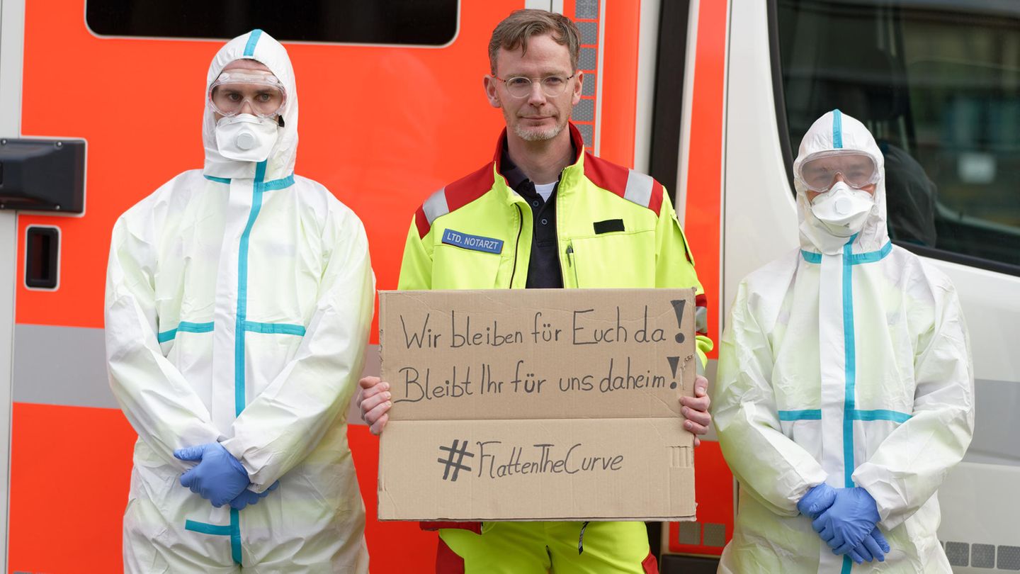 #FlattenTheCurve: Ein Notarzt hält, eingerahmt von zwei Rettungssanitätern in Infektionsschutzkleidung, ein Schild mit der Aufschrift "Wir bleiben für euch da! Bleibt für uns daheim!".
