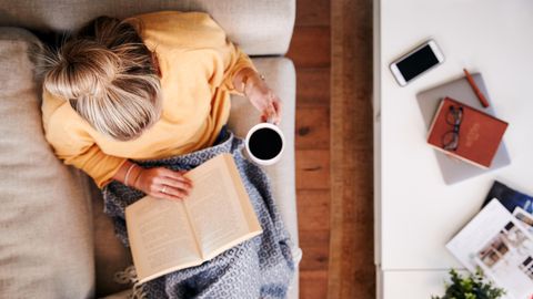 Frau sitzt mit einem Buch und einer Tasse Kaffee im Bett