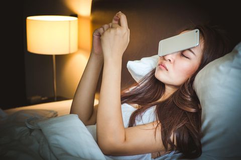 Eine Frau liegt mit einem Smartphone, das ihr aus den Händen auf das Gesicht gefallen ist, im Bett
