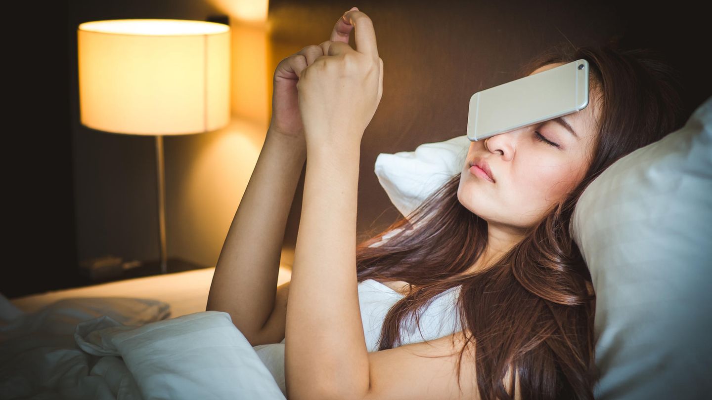 Eine Frau liegt mit einem Smartphone, das ihr aus den Händen auf das Gesicht gefallen ist, im Bett