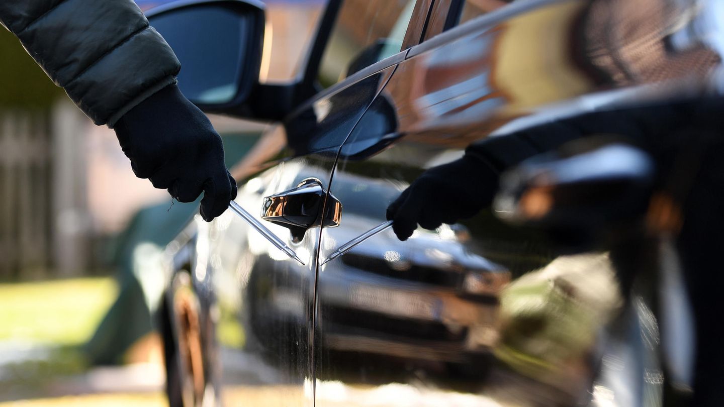 Ein Mann steht neben einem Auto und hält einen Schraubenzieher in der Hand. Er trägt schwarze Handschuhe
