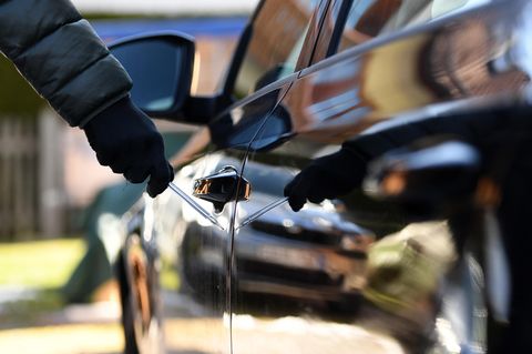 Ein Mann steht neben einem Auto und hält einen Schraubenzieher in der Hand. Er trägt schwarze Handschuhe