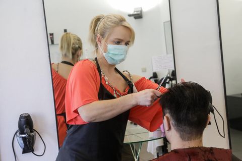 Eine blonde Friseurin in rotem T-Shirt trägt einen Mundschutz, während sie einem Mann die braunen Haare schneidet