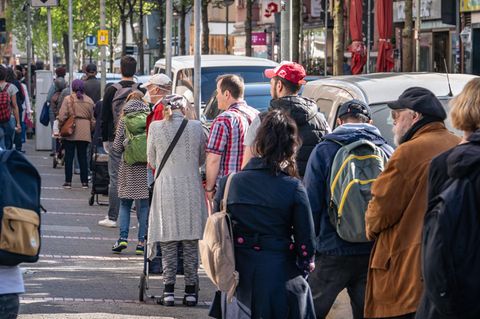 Männer und Frauen unterschiedlichen Alters stehen bei Sonne auf einem Fußweg Schlange
