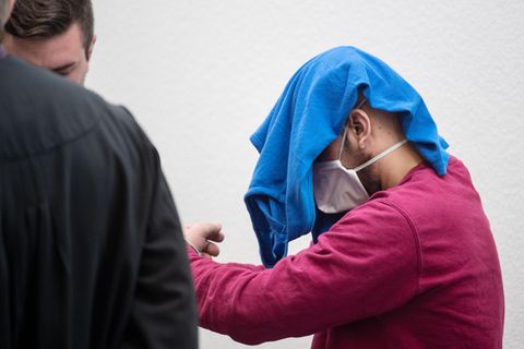 Der Angeklagte betritt in Handschellen, mit einem blauen T-Shirt und Mundschutz den Gerichtssaal
