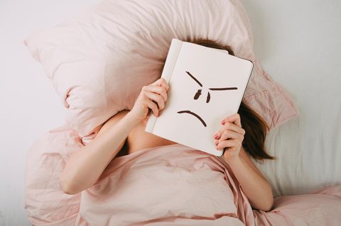 Frau liegt im Bett und hält ein Buch vor ihr Gesicht, auf dem sie ein wütend schauendes Smiley gemalt hat
