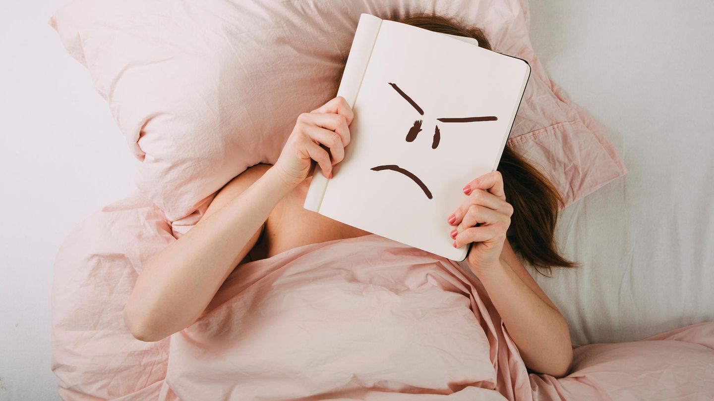 Frau liegt im Bett und hält ein Buch vor ihr Gesicht, auf dem sie ein wütend schauendes Smiley gemalt hat