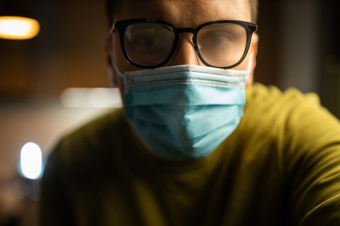 Ein Mann mit gelben Pulli trägt einen blauen medizinischen Mundschutz. Seine Brille ist beschlagen