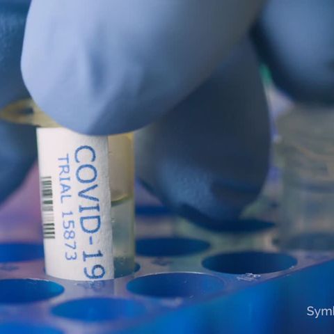 Finger in einem blauen Handschug greifen eine Plastik-Ampulle mit "Covid-19"-Beschriftung aus einem blauen Halter