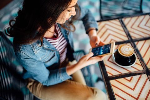 Frau sitzt mit Handy in der Hand und einer Kaffeetasse an einem Tisch