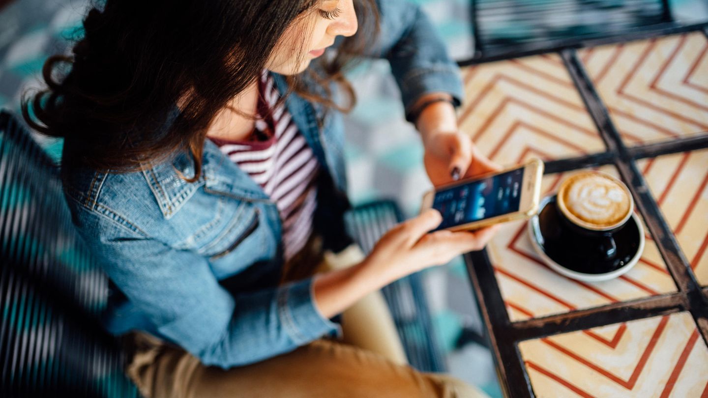 Frau sitzt mit Handy in der Hand und einer Kaffeetasse an einem Tisch