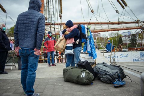 Mutter und Tochter umarmen sich zur Begrüßung. Im Hintergrund ist ein hölzerner Mast eines Segelschiffs zu sehen