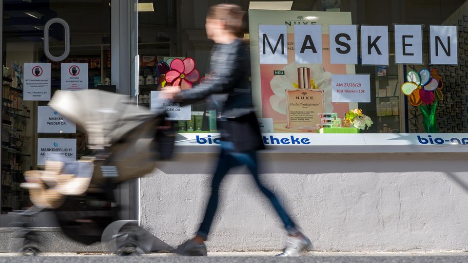 Eine Frau schiebt einen Kinderwagen an einer Apotheke vorbei, die Mundschutzmasken anbietet