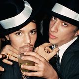 Eine Frau und ein Mann posieren in Nadelstreifenanzügen mit Hüten, Weingläsern und Zigarre