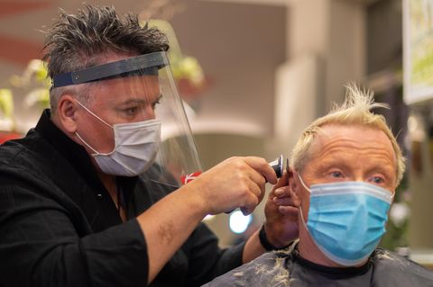 Ein Frisuer mit Atemmaske und Geischtsvisir rasiert einem Kunden mit Mundschutz die Haare am Hinterkopf