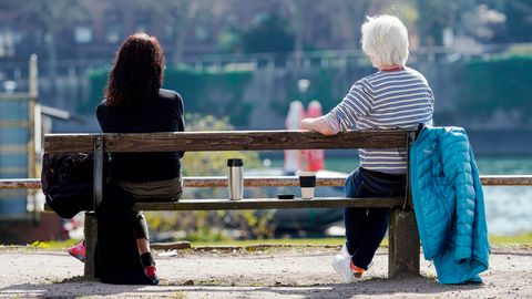 Auf einer Parkbank sitzen zwei Frauen. Rechts eine Weißhaarige, links eine Brünette. In der Lücke zwischen ihnen Kaffeebecher